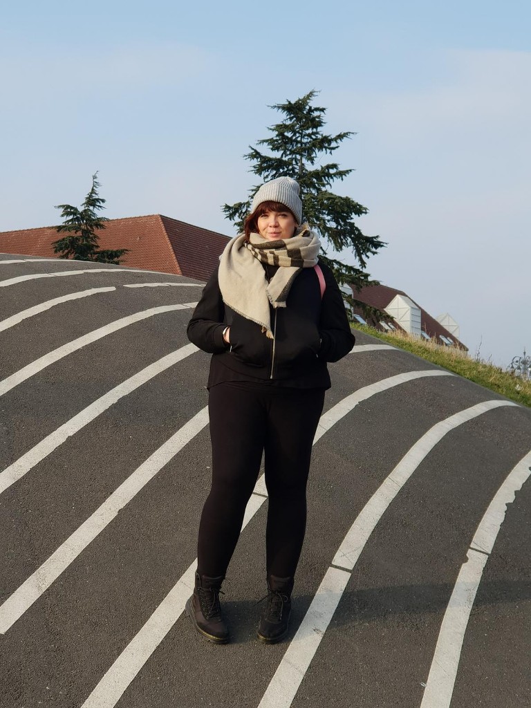 Lesley standing on curved striped ground wearing a beanie hat, scarf and dark jacket, with trees and rooftops behind her