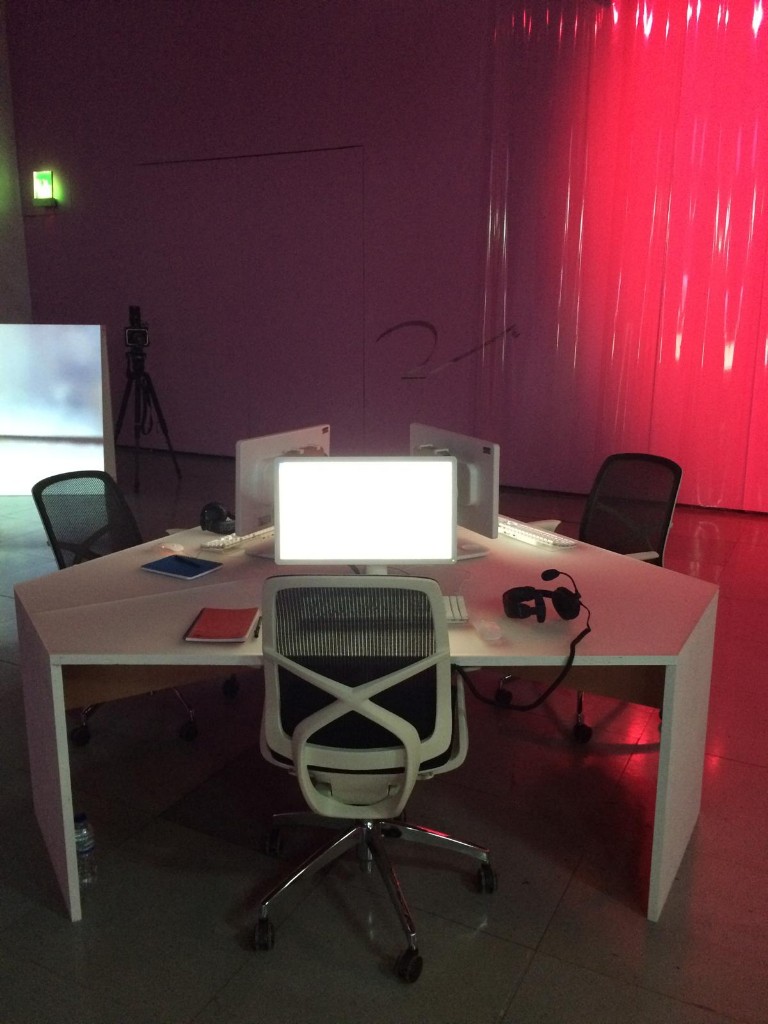 Gallery installation showing a cluster of white desks with computer monitors and VR headsets, bathed in red light from translucent plastic curtains